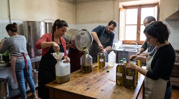 Varias personas rellenan botellas y bidones plásticos con aceite de oliva usando un barril metálico y embudos en una cocina.