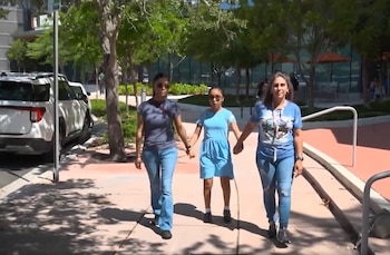 Três pessoas, duas mulheres mais velhas e uma menina de vestido azul, caminham de mãos dadas em uma calçada pavimentada ao lado de um caminho arborizado sob a luz do sol.
