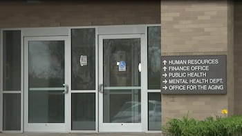 Entrada de edificio con puertas de vidrio y símbolo de accesibilidad. Un cartel en la pared de ladrillo dirige a departamentos como Recursos Humanos y Salud Pública