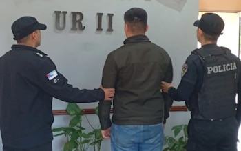 Dos policías escoltan a un hombre vestido de civil. Los agentes visten uniforme oscuro, gorra y uno tiene chaleco. Fondo con letras 'UR II' y plantas