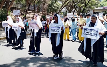 Grupo de mujeres, algunas con velos blancos y túnicas oscuras, marchan en una calle arbolada sosteniendo pancartas con mensajes de protesta
