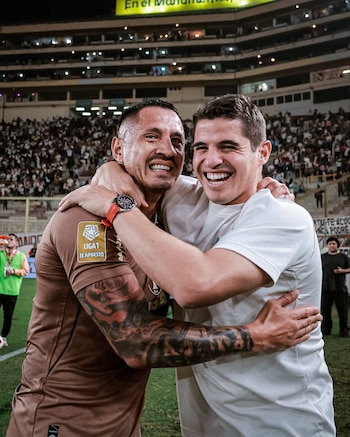 Dos hombres sonríen y se abrazan en un campo de fútbol. Uno con camiseta marrón y tatuajes, el otro con camiseta blanca. Fondo de estadio con público