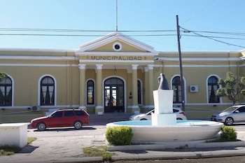 Edificio amarillo con la inscripción Municipalidad, columnas en el pórtico, una fuente blanca en primer plano y tres automóviles