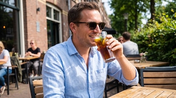 Hombre sonriente con camisa celeste y gafas de sol bebe té helado con limón y menta en vaso de vidrio, sentado en una mesa de madera al aire libre.
