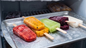 Cinco paletas de hielo de distintos colores con palitos de madera organizadas sobre una bandeja metálica dentro de un congelador.