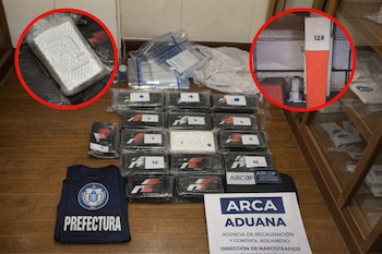 Vista aérea de múltiples paquetes de cocaína envueltos, un chaleco azul con el logo de Prefectura y un cartel blanco de ARCA Aduana, sobre un suelo de madera.