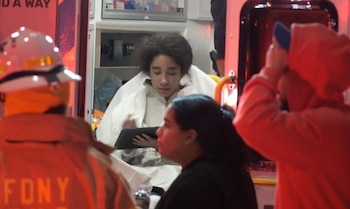 Joven con cabello rizado sentado en el interior de una ambulancia sosteniendo una tableta, con un bombero del FDNY de espaldas y otras personas en primer plano