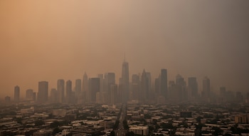 Vista panorámica de una gran ciudad con rascacielos y edificios, una avenida con vehículos, bajo una capa de humo espeso que tiñe el cielo de naranja.