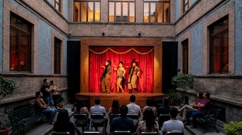 Vista aérea de un patio interior con un escenario iluminado donde tres artistas de cabaret actúan frente a una audiencia sentada en sillas y bancos.