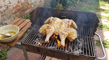 Un pollo entero en mariposa se asa sobre una parrilla con carbón y llamas. Junto a él, un cuenco blanco con aderezo y un pincel de cocina.