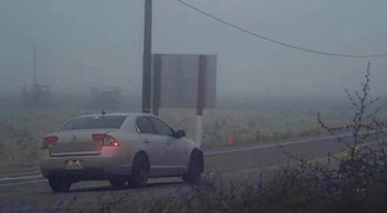 Un automóvil gris estacionado a un costado de una carretera asfaltada bajo la niebla, junto a un poste de madera y una señalización