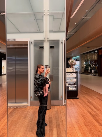 Uma mulher com um bebê nos braços tira uma selfie em frente ao espelho. Você pode ver os elevadores e corredores do shopping center com lojas