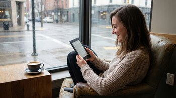 Mujer sonriente leyendo un Amazon Kindle en un sillón junto a una ventana. Hay una taza de café en una mesa de madera. Fuera, una calle urbana mojada por la lluvia.