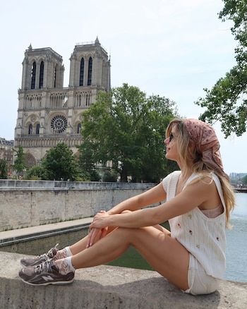 Laurita Fernández sentada en un muro de piedra con la Catedral de Notre Dame de París y un río al fondo, bajo un cielo claro con árboles