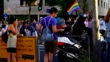 Calle con personas, bandera arcoíris, motocicleta blanca y negra, hombre con casco, hombre con camiseta morada y mochila, mujer, cartel amarillo con texto