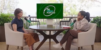 Oprah Winfrey y una mujer sentadas frente a una mesa redonda de madera, grabando un podcast. Micrófonos, vasos y el logo 'The Oprah Podcast' son visibles. Fondo de jardín