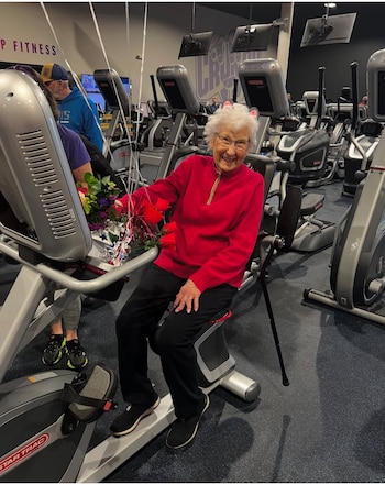 Una mujer con suéter rojo y lentes permanece sentada en una bicicleta fija de un gimnasio, con flores, globos y un bastón a su lado