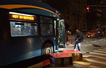 Un autobús de la MTA azul y amarillo en la calle por la noche, con la puerta abierta, junto a restos de un comedor y un vehículo naranja al fondo