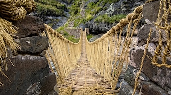 Un puente de cuerdas tejidas y madera que cruza un cañón entre pilares de piedra con un sendero de escalones al fondo.