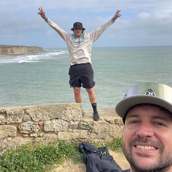 Selfie de un hombre sonriendo en primer plano, con otro hombre con sombrero y brazos levantados de pie en un muro de piedra junto al océano y acantilados al fondo