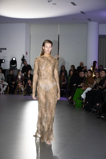 Una modelo camina por la pasarela con un vestido largo de malla dorada brillante ante el público sentado