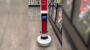 Robot vertical blanco y rojo con un cartel que dice "Hi, I'm Tally! I check shelf inventory!" frente a congeladores.
