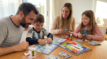 Una familia de cuatro personas sentada alrededor de una mesa de madera, pegando figuritas del álbum del Mundial FIFA 2026. Hay un padre, una madre y dos niños.