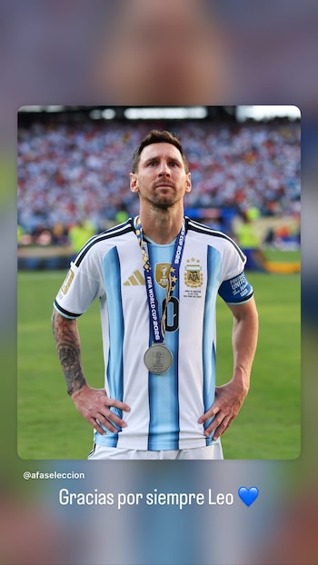 Hombre, tez blanca, pelo corto castaño, barba corta, camiseta celeste y blanca selección argentina. Medalla plateada con cinta azul al cuello. Parche en brazo. Brazos en jarras. Fondo de cancha de fútbol con público borroso