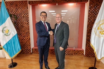 Dos hombres con traje de saco y corbata se dan la mano. Detrás, una bandera de Guatemala y otra blanca con el escudo del Ministerio de Economía