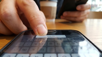 Primer plano enfocado en dedo presionando barra espaciadora de teclado de celular. Se observa taza de café y otra mano borrosa al fondo.