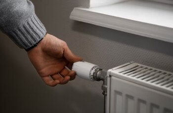 Primer plano de una mano girando el regulador blanco de un radiador metálico de color claro, situado junto a una pared gris y un alféizar blanco