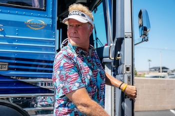 Hombre rubio con perilla, vistiendo visera blanca 'HAWK' y camisa azul de águilas, parado junto a la puerta de un autobús azul bajo el cielo claro