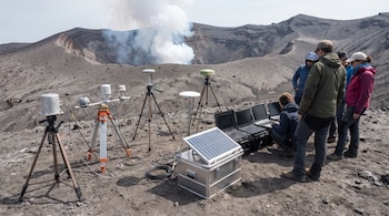 Un volcán con humo en el cráter, equipos científicos con trípodes, un panel solar, varias computadoras portátiles y un grupo de personas observando.