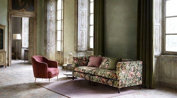 Salón de estilo clásico con paredes verdes, dos ventanas, cortinas, sofá estampado floral, dos cojines, sillón rosa, mesa auxiliar y una alfombra.