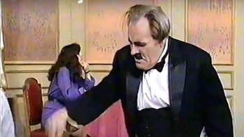 Primer plano de Tandarica, un hombre con bigote, vestido con esmoquin negro y camisa blanca. Una mujer con cabello oscuro está sentada de espaldas en el fondo