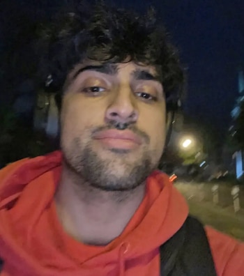 Fotografía en primer plano de un joven con cabello rizado, barba corta, auriculares negros y sudadera roja con capucha de noche en la calle