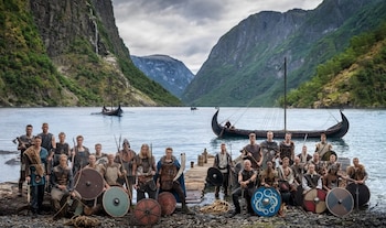 Grupo de hombres vestidos como vikingos con escudos y armas posan a la orilla de un fiordo noruego, con barcos vikingos y montañas de fondo