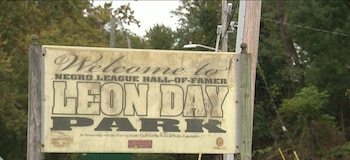 Cartel de entrada al parque Leon Day en Baltimore con letras negras sobre un fondo blanco desgastado y vegetación detrás