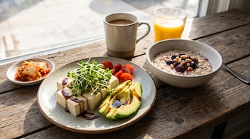 Plato con dados de tofu, brotes germinados, aguacate y tomates cherry junto a un tazón de avena, kimchi, una taza de café y un vaso de jugo.