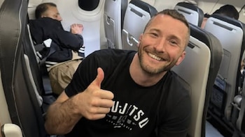 Un hombre barbudo, sonriendo y haciendo un gesto de aprobación con el pulgar arriba, dentro de la cabina de un avión. Viste una camiseta negra y se ven asientos y pasajeros detrás