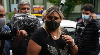 Una mujer de cabello rubio con mascarilla de camuflaje y camiseta negra con pedrería, flanqueada por hombres que sostienen cámaras y un micrófono.