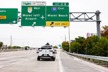 Un vehículo autónomo blanco de Waymo con sensores en el techo circula por una autopista de varias pistas bajo señalización vial para el Aeropuerto de Miami y Miami Beach