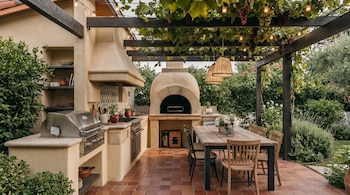 Cozinha externa com churrasqueira, forno de pizza, mesa e cadeiras de madeira, piso frio, pergolado com trepadeiras e luminárias suspensas, plantas e arbustos.
