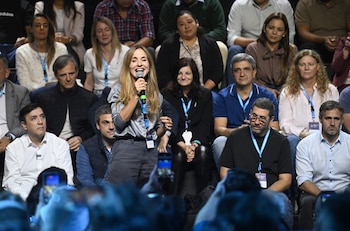 Una mujer con cabello largo y rubio, vestida con blusa plateada y pantalones oscuros, sonríe mientras habla por un micrófono azul frente a una audiencia variada