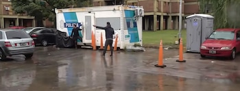 Vista exterior de una unidad policial móvil y dos hombres en un área húmeda, con autos estacionados y conos de tráfico naranjas