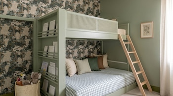 Literas de color verde salvia con detalle de ratán y escalera de madera en una habitación con papel tapiz de bosque y paredes verdes; incluye estantería y juguetes.