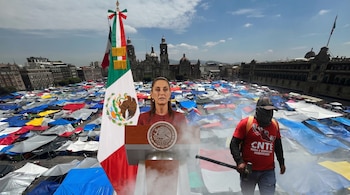 Claudia Sheinbaum en un podio con la bandera de México, frente a un Zócalo lleno de tiendas de campaña de colores, junto a un manifestante de la CNTE con un martillo.