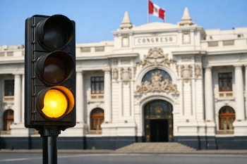 Un semáforo de tres luces, con la luz amarilla inferior encendida, se sitúa frente al edificio del Congreso de la República del Perú con una bandera peruana.