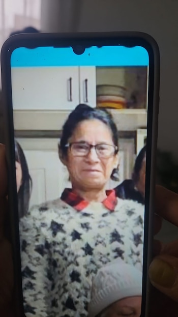 Una mujer con cabello oscuro, gafas y un suéter estampado se muestra en la pantalla de un teléfono inteligente sostenido por una mano