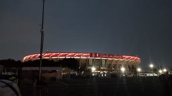 Vista nocturna del Estadio Ciudad de México iluminado con luces rojas en su exterior bajo un cielo oscuro, con vallas y postes en primer plano